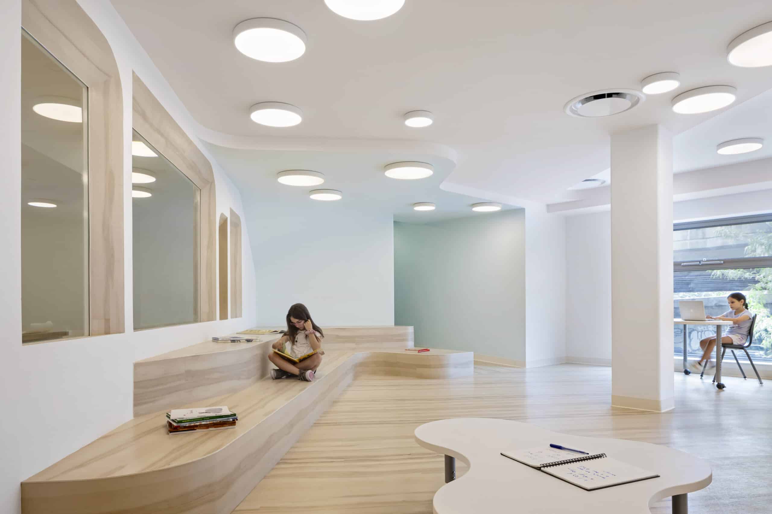 Children studying in a simplistic and modern classroom-like space, featuring wooden platforms as part of the furniture.