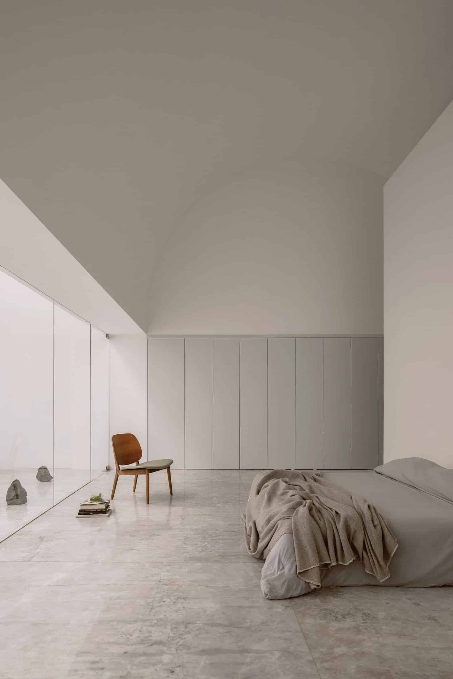White interior room with an arched ceiling, built-in white wardrobes, a bed with grey bedding, a wooden chair and sliding glass doors