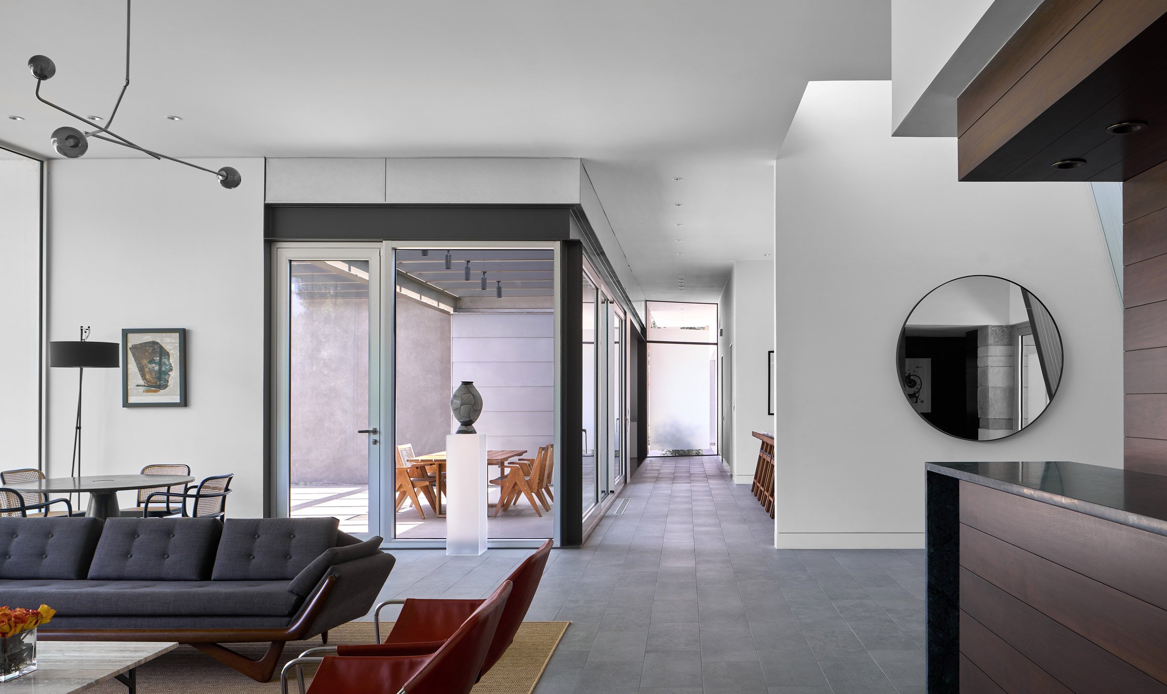 Open-plan living space with white walls, porcelain floor tiles, a grey sofa and corner glazing opening to an outdoor patio