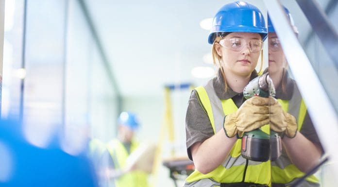 a teenage female carpenter is installing handrails in a modern office refurbishment. IN the background a co-worker can be seen holding plans . They are wearing safety workwear. Representing building control apprenticeships