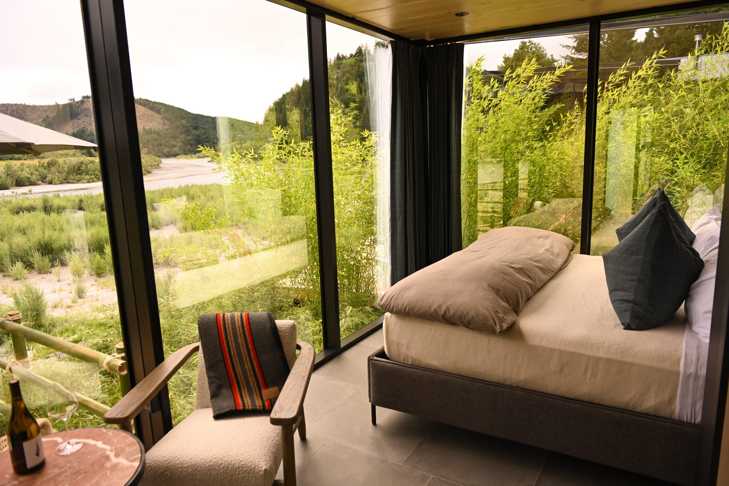 Glass cabin interior with a double bed, side table and chair overlooking a river