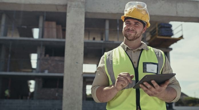 A building contractor using a digital tablet while working on a construction site. A young man browsing online with a smart device while working on an architecture project, representing utilising both Newforma and Procore