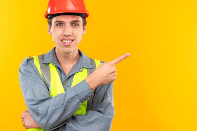 smiling young builder man in uniform points at side isolated on yellow background with copy space
